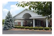 Covered front entrance of a brick senior living facility with a porte-cochere, shrubs, and a pine tree.