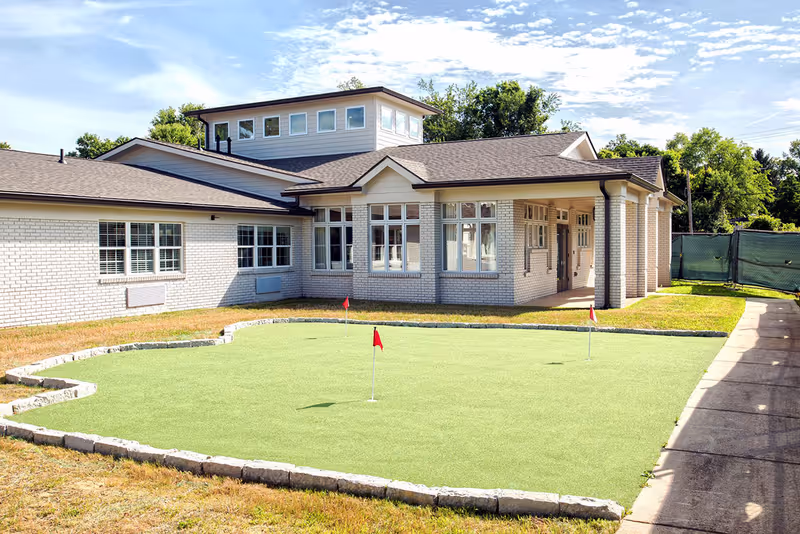 Exterior view of a single-story brick senior living building with a small putting green and red flags in the foreground.