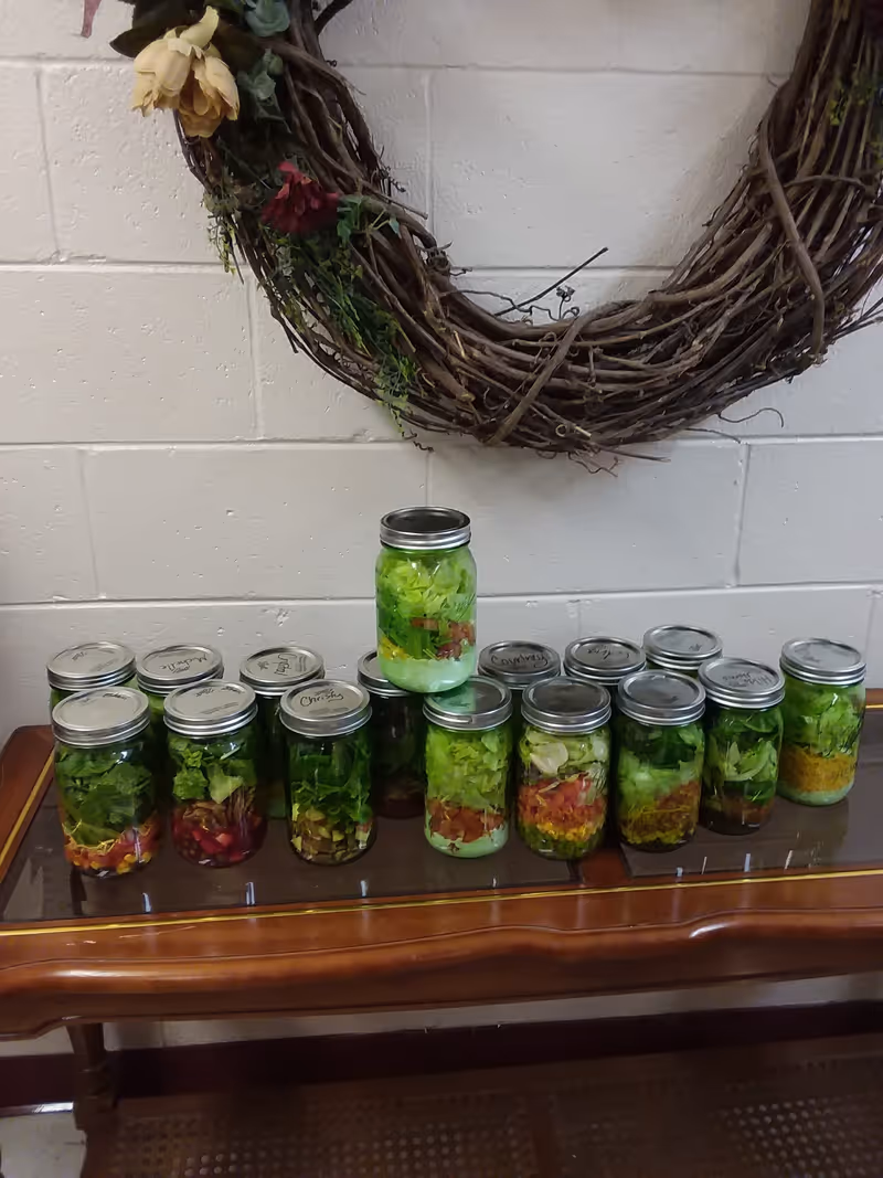 Row of mason jars filled with layered greens on a wooden bench beneath a wreath.