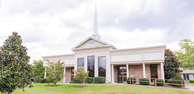 Exterior view of a single-story building with a tall white steeple, large windows, brick and beige siding, surrounded by green grass, bushes, and trees under a cloudy sky.