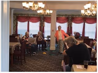 A group of elderly people sitting and standing in a dining room with tables covered in white tablecloths. The room has large windows with red curtains and chandeliers hanging from the ceiling. One person is standing near a white pillar, while others are seated at tables.