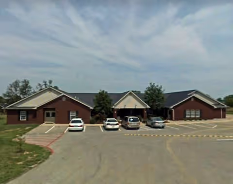 Single-story brick assisted living building with a central covered entrance and a small parking lot with several cars.
