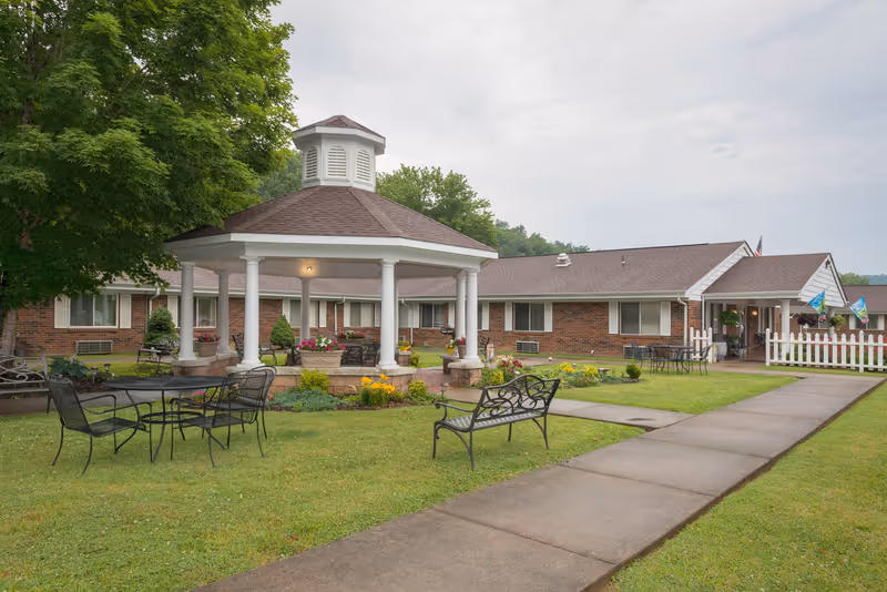 Outdoor view of Laurel Creek Health Care Center featuring a brick building with white trim, a white gazebo with a brown roof in the center of a grassy area, black metal benches and tables, flower beds, and a paved walkway leading to the entrance.