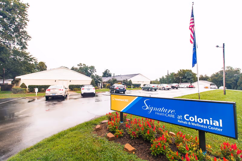 Exterior view of Signature HealthCARE at Colonial Rehab & Wellness Center with its blue sign, flagpole, parking lot and facility buildings on a wet day.