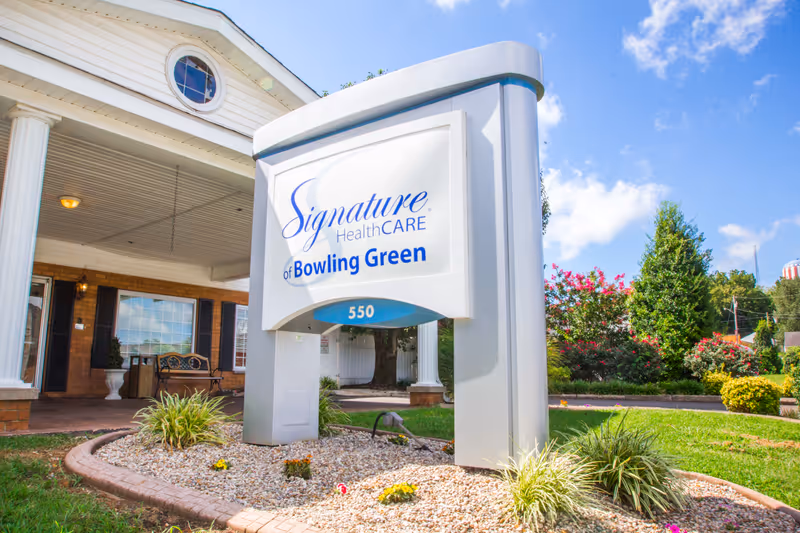 Outdoor view of the entrance to Signature HealthCARE of Bowling Green, featuring a large white and blue sign with the facility name and address number 550, surrounded by landscaping with grass, bushes, and flowers under a partly cloudy blue sky.