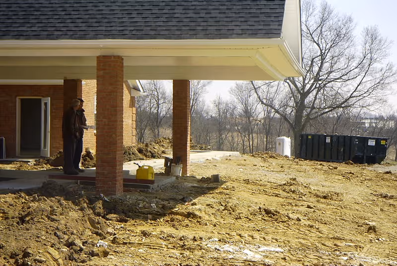 Two people stand near a brick-columned building entrance at a construction site.