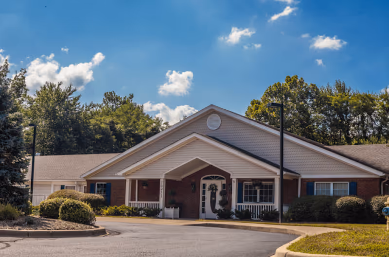 Front entrance of a one-story brick senior living building with a covered portico, landscaped shrubs, and trees under a blue sky.