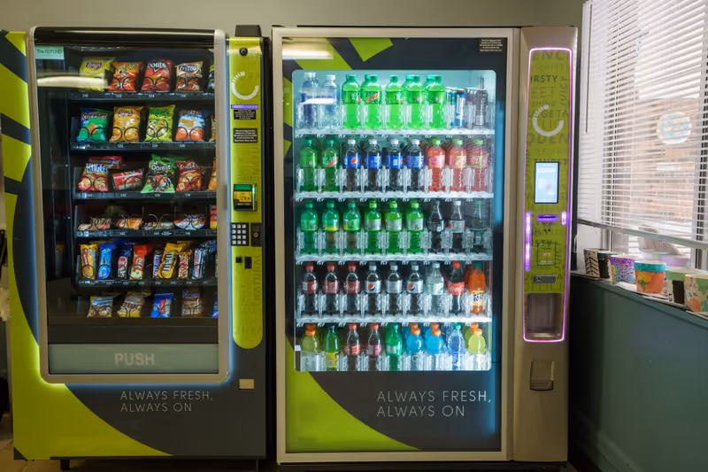 Two vending machines side by side inside a room. The left machine contains various snack bags including chips and candy bars, while the right machine is stocked with bottled beverages such as water, soda, and sports drinks. The machines have green and black accents and the phrase 'ALWAYS FRESH, ALWAYS ON' is visible on both. There is a window with blinds and some colorful cups on the windowsill to the right.