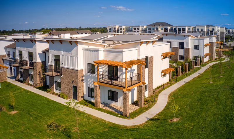 Modern multi-unit residential building with balconies, landscaped lawn and a curved walking path under a blue sky.