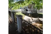 A sunny outdoor garden area with a white gazebo surrounded by a white picket fence. There is a black metal gate and fence in the foreground, and three ducks walking on a paved path leading to the gazebo. Trees provide shade over the area.