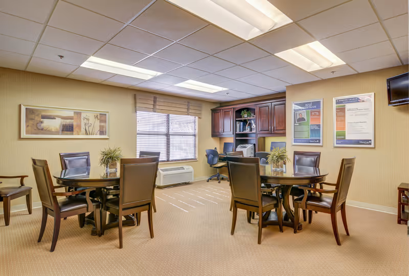 A well-lit room with two round tables, each surrounded by four brown leather chairs. There are two plants in glass vases on the tables. A window with blinds and a beige valance is on the back wall. To the right, there is a wooden cabinet with office supplies and a printer, along with two framed posters on the wall and a mounted flat-screen TV. The room has beige walls and carpet, and a drop ceiling with fluorescent lights.