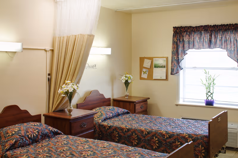 A bedroom with two single beds featuring floral patterned bedspreads. Each bed has a wooden headboard and a wooden nightstand with a vase of flowers. A beige curtain is partially drawn between the beds. There is a window with blinds and a floral valance, and a small plant in a purple pot on the windowsill. A bulletin board with papers and a calendar is mounted on the wall.