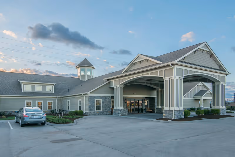 Exterior view of The Lantern at Morning Pointe Alzheimer’s Center of Excellence in Lexington showing a large covered entrance with columns, a stone and siding facade, a parked car, and a partly cloudy sky at dusk.