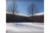 Snow-covered courtyard in front of a two-story brick building with leafless trees under a clear blue sky.