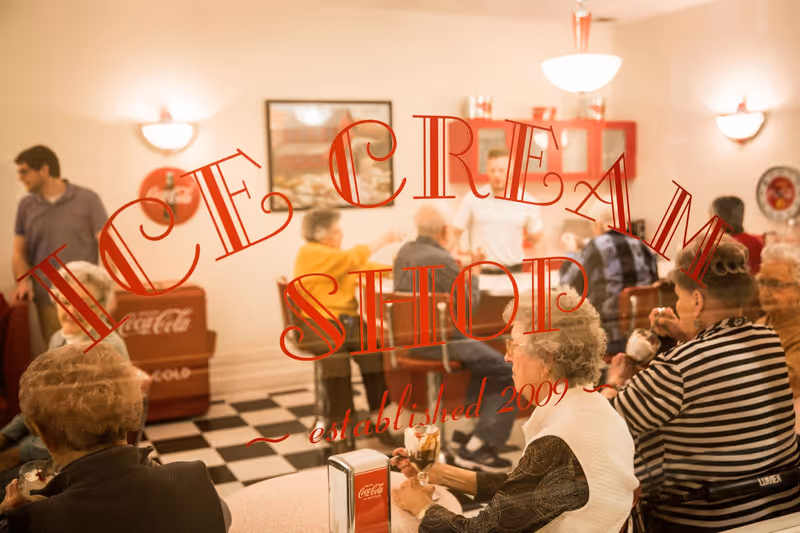View through a glass window with red lettering that reads 'ICE CREAM SHOP established 2009' showing elderly people sitting and enjoying ice cream sundaes in a retro-style ice cream parlor with checkered floor and Coca-Cola decor.
