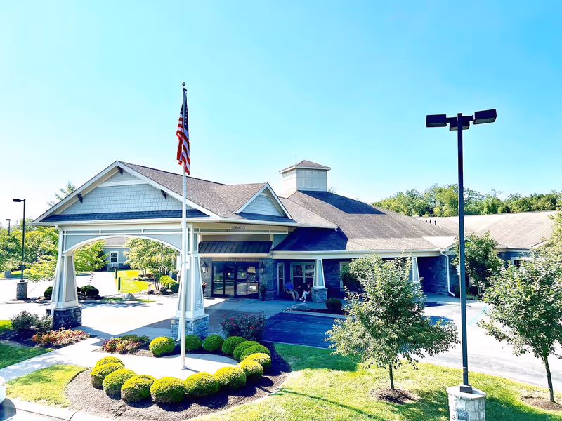 Exterior view of Morning Pointe of Lexington-East facility showing the main entrance with a covered driveway, an American flag on a flagpole, landscaped bushes and trees, and a clear blue sky.