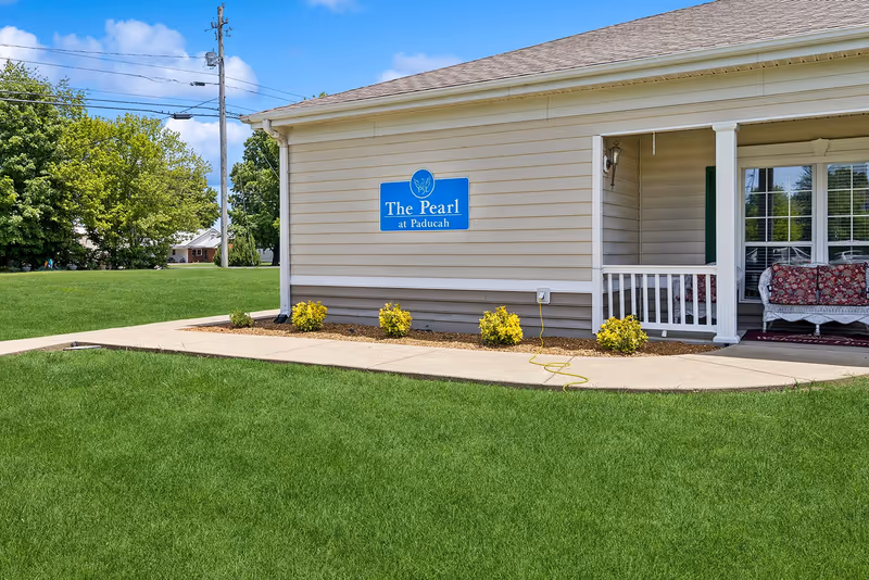 Exterior view of a single-story building with beige siding and a small covered porch featuring white railings and floral cushioned seating. A blue sign on the building reads 'The Pearl at Paducah'. The building is surrounded by a well-maintained green lawn and small shrubs, with trees and a clear blue sky in the background.