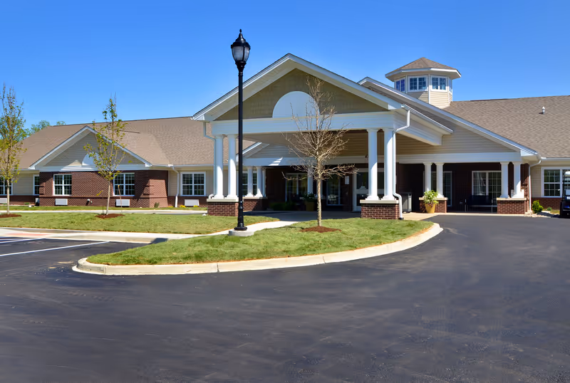 Exterior view of a single-story senior living facility building with a covered entrance supported by white columns, a small landscaped area with young trees and grass, and a clear blue sky.