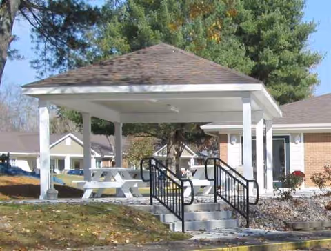 Outdoor covered pavilion with white picnic table, stairs, and brick building in the background.