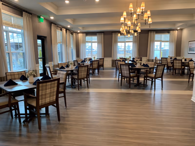 A spacious dining room with multiple wooden tables and plaid cushioned chairs. Each table is set with dark blue folded napkins and small white flower arrangements. Large windows with white curtains allow natural light to fill the room. A modern chandelier with multiple lampshades hangs from the ceiling.