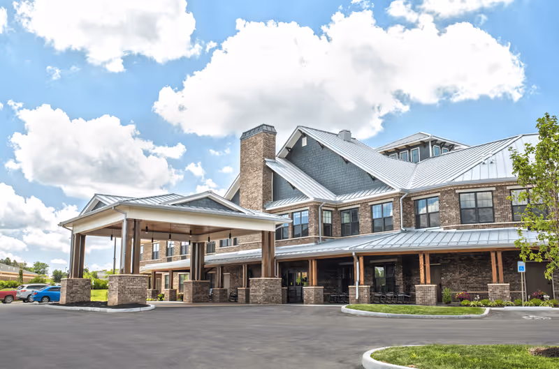 Exterior view of a large brick senior living facility building with a covered entrance, multiple windows, and a metal roof under a partly cloudy sky. There are a few parked cars and some landscaping with grass and small trees around the building.