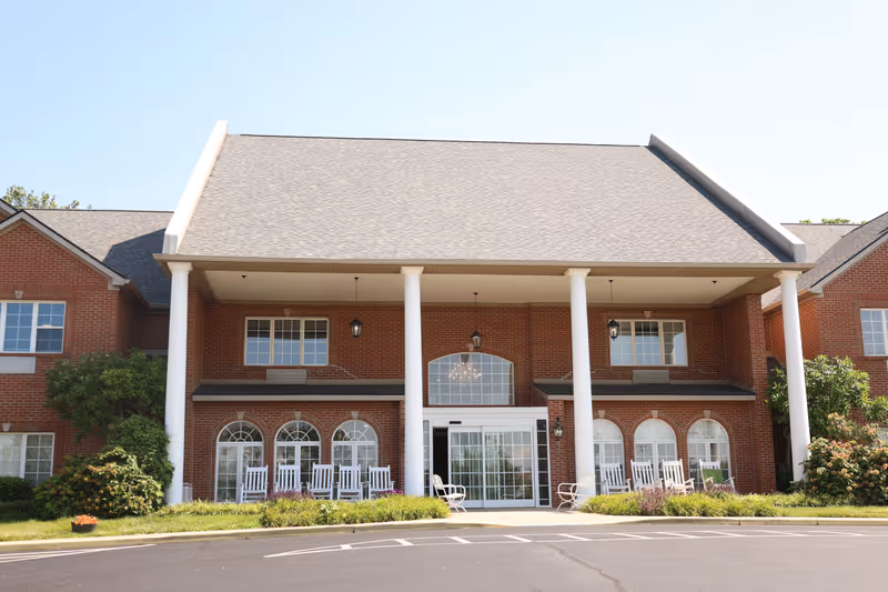 Front exterior of a two-story brick senior living building with white columns, arched windows, and rocking chairs on the covered entrance porch.