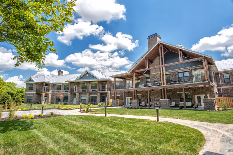 Exterior view of Dominion Senior Living of Florence, showing a large two-story brick building with balconies and a covered patio area. The building is surrounded by a well-maintained lawn, landscaped garden beds, and a paved walkway under a partly cloudy blue sky.