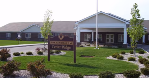Exterior view of Cedar Ridge Health Campus showing a single-story building with a covered entrance, a flagpole, and a landscaped lawn with small trees and shrubs. A sign in front reads 'The Residence at Cedar Ridge'.