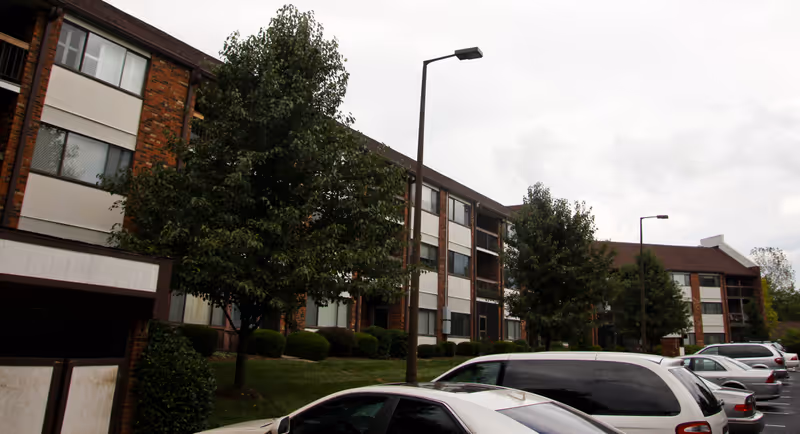 Exterior view of a multi-story senior living facility building with brick and white paneling, surrounded by trees and parked cars in the parking lot on a cloudy day.