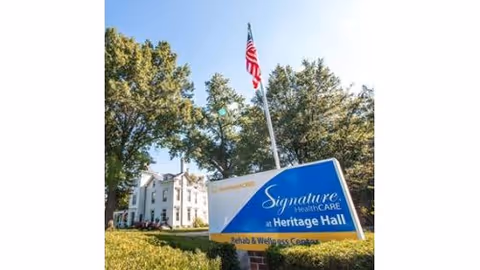 Front view of Signature HealthCARE at Heritage Hall sign with flag and trees