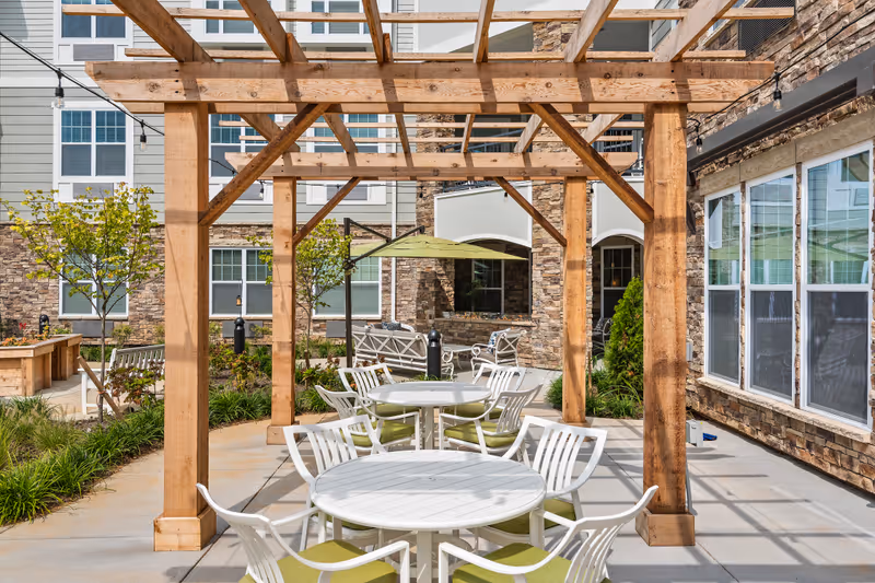 Outdoor seating area at The Ashton on Dorsey featuring white round tables with green cushioned chairs under a wooden pergola. The area is surrounded by plants, small trees, and stone building walls with large windows.