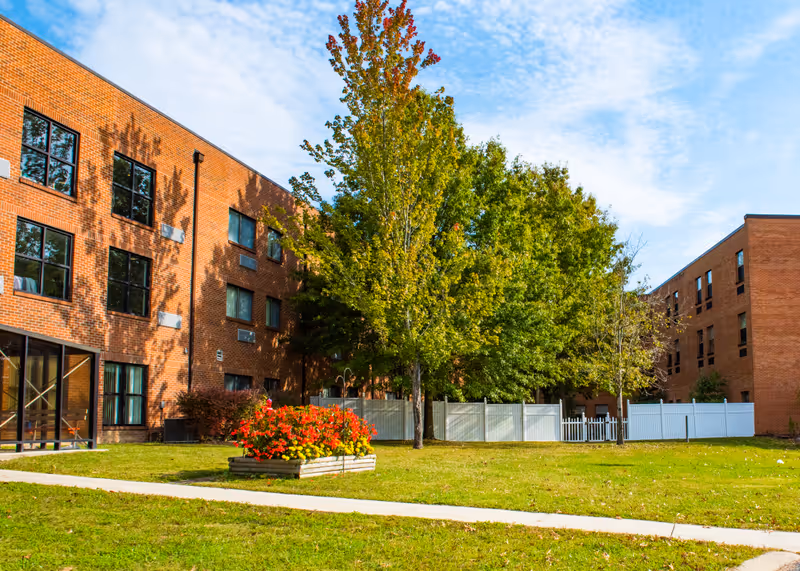 Outdoor view of a senior living facility with two brick buildings on either side, a green lawn with a flower bed of red and yellow flowers, several trees with green and some autumn-colored leaves, and a white fence in the background under a partly cloudy sky.