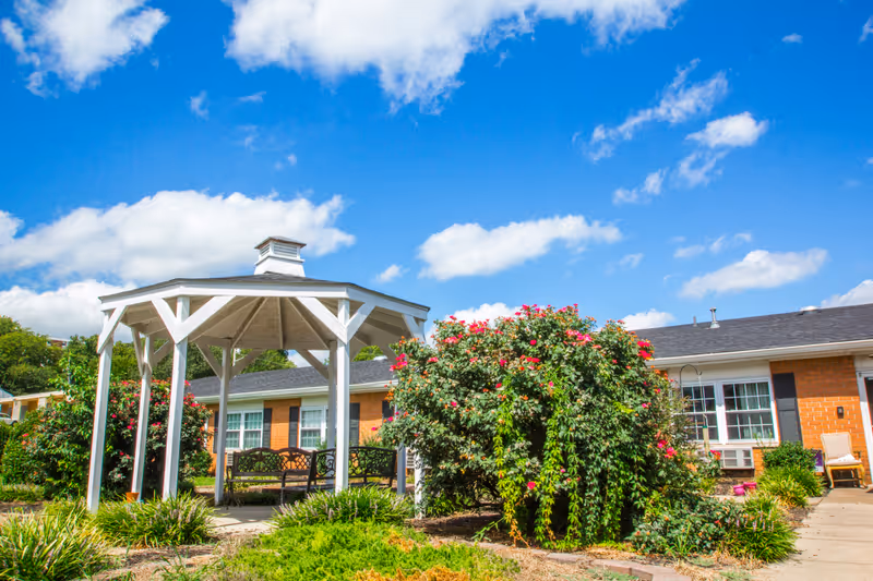 Outdoor garden area at a senior living facility with a white gazebo, benches underneath, blooming bushes with pink flowers, and a brick building in the background under a blue sky with scattered clouds.