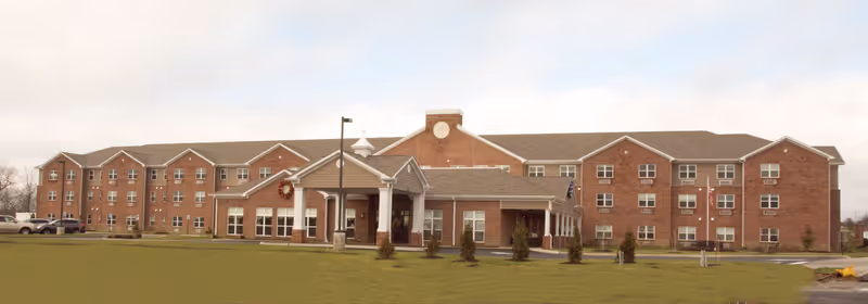 Exterior view of a large, three-story brick senior living facility building with multiple windows, a covered entrance with white columns, a wreath decoration, and a flagpole with an American flag in front. The building is surrounded by a grassy area and a few small trees under a partly cloudy sky.