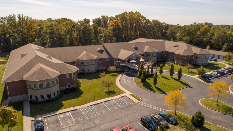 Aerial view of Magnolia Springs East Louisville, a large senior living facility with a U-shaped building surrounded by green lawns, trees, and a parking lot with several cars. The building has multiple windows and a covered entrance with a circular driveway.