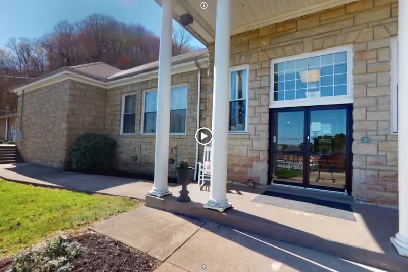 Exterior view of a building entrance with stone walls, two white columns, a glass double door, and windows. There is a small porch area with a rocking chair and potted plants, and a grassy area with a sidewalk leading to the entrance.
