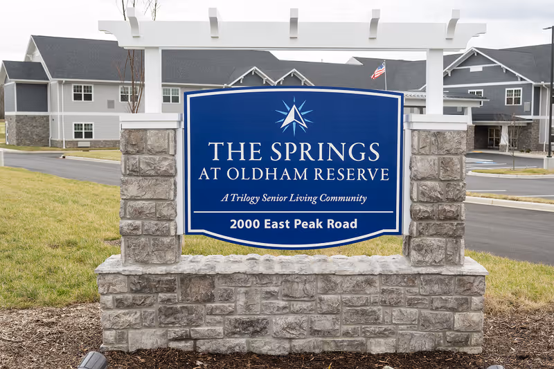 Blue entrance sign for The Springs at Oldham Reserve (2000 East Peak Road) in front of the senior living building.
