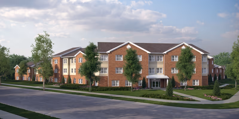 Exterior view of a three-story senior living residence building with a combination of red brick and beige siding. The building is surrounded by green lawns, trees, and landscaped flower beds under a partly cloudy sky.