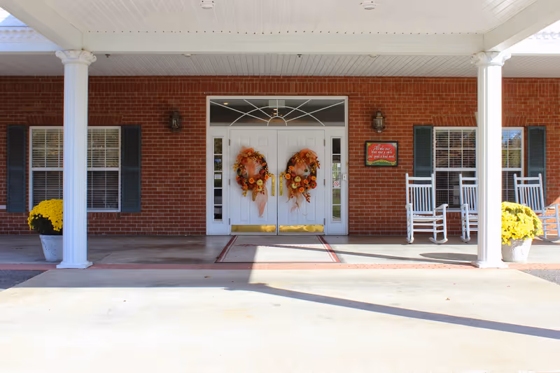 Front entrance of Oak Ridge Senior Living Community featuring double white doors decorated with autumn wreaths, flanked by two white columns. There are two windows with dark shutters on either side of the doors, white rocking chairs on the right side, and large pots of yellow flowers on both sides of the entrance. A red sign with white text is mounted on the brick wall to the right of the doors.