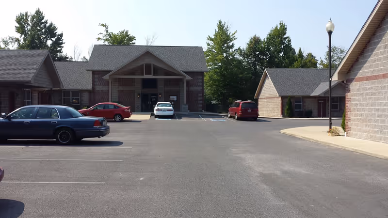 Parking lot with several parked cars in front of a brick and stone building with a peaked roof and entrance canopy, surrounded by trees under a clear sky.