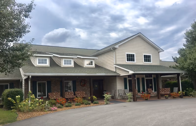 Exterior view of a single-story assisted living facility building with a covered porch area featuring several chairs and floral-patterned cushions. The building has beige siding, a green roof, multiple windows, and landscaped flower beds in front. The sky is partly cloudy.