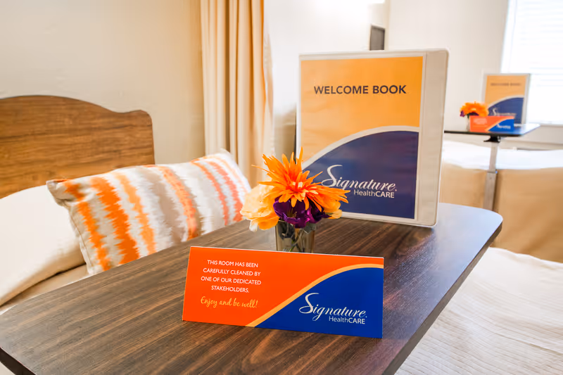 A close-up view of a wooden table in a bedroom with a colorful pillow on a bed in the background. On the table, there is a small vase with orange and purple flowers, a welcome book with the Signature HealthCARE logo, and a card indicating the room has been carefully cleaned by dedicated stakeholders.