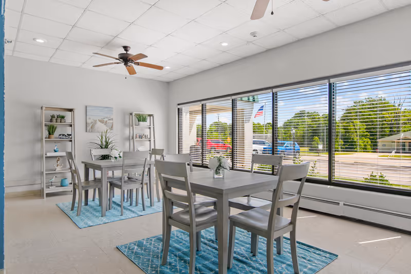 A bright and clean dining area with two wooden tables and eight matching chairs placed on blue patterned rugs. The room has large windows with blinds, allowing natural light to fill the space and offering a view of parked cars and greenery outside. Two ceiling fans are mounted on the white tiled ceiling, and two shelves with decorative plants and items are against the wall.