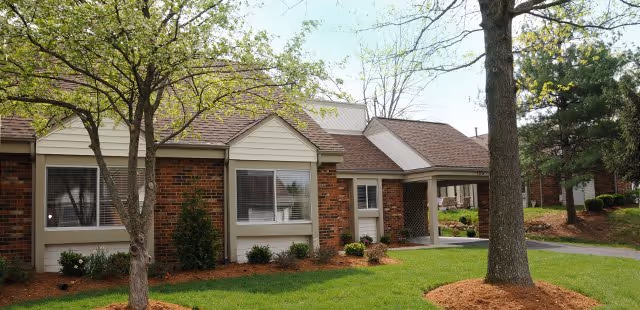 Front exterior of a single-story brick senior living building with a covered entrance, lawn and trees.