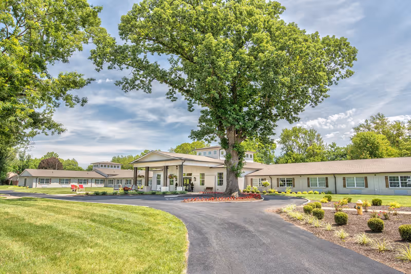 Exterior view of Dominion Senior Living of Louisville, showing a single-story building with a covered entrance, surrounded by well-maintained landscaping including a large tree, bushes, and green lawn under a partly cloudy sky.