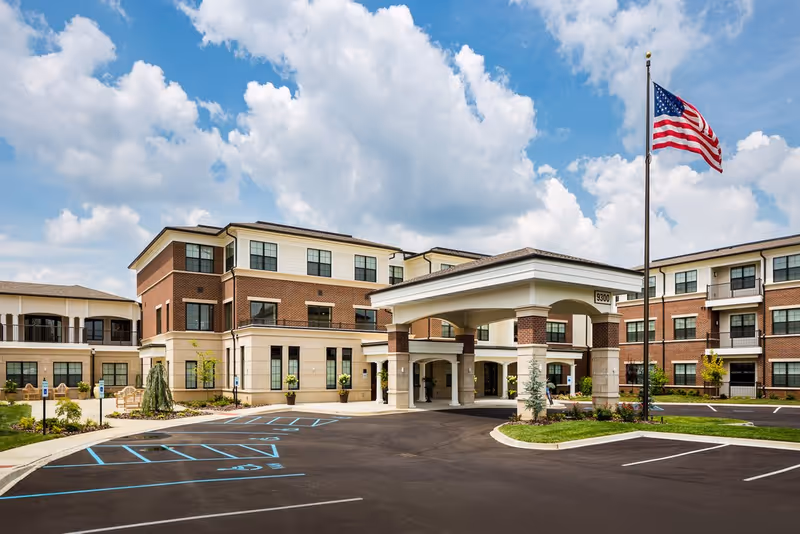 Exterior view of a three-story senior living facility with a covered entrance, brick and beige facade, multiple windows, an American flag on a flagpole, and a parking lot with marked spaces including handicapped spots.