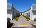 Two single-story white buildings facing each other with a central paved walkway, low shrubs, and a clear blue sky.
