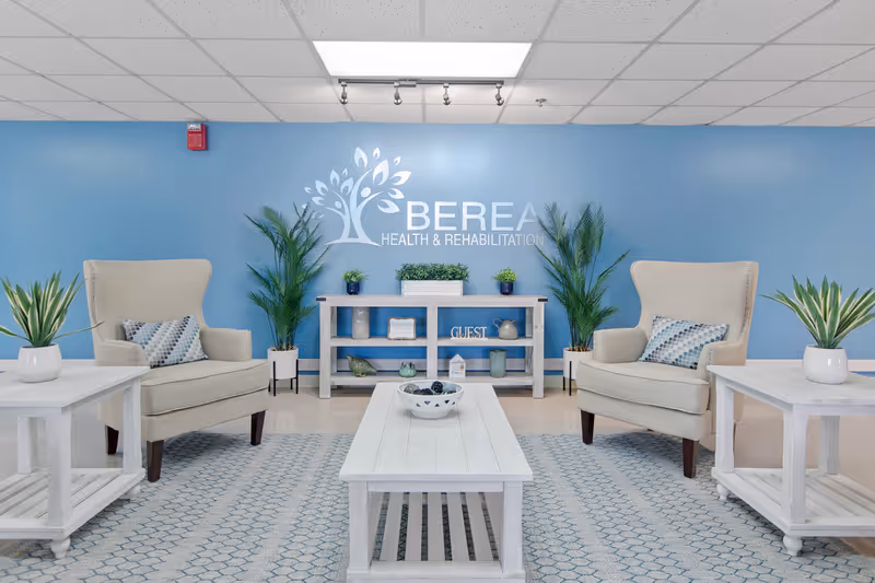 A bright and welcoming lounge area with two beige armchairs, white wooden tables, and a white coffee table on a patterned rug. The background features a blue wall with the Berea Health & Rehabilitation logo, flanked by green potted plants and decorative items on a white console table.