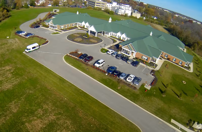 Aerial view of Daisy Hill Senior Living facility showing a large single-story building with green roofs, surrounded by parking lots with several cars, landscaped areas, and green lawns under a clear blue sky.