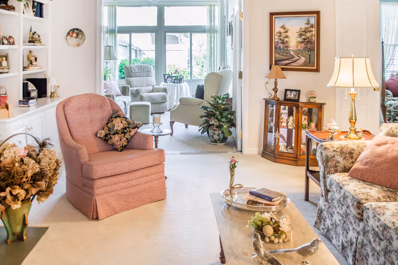 Cozy living room with pink armchair, floral sofa, lamps, and sunlit enclosed porch.